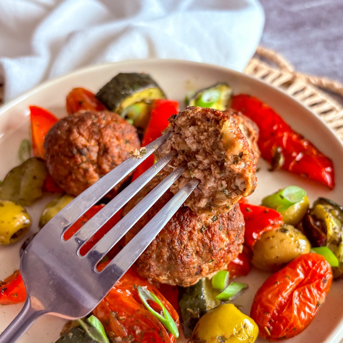 Baked Mediterranean meatballs being eaten, a fork lifting half a meatball from a cream bowl of roasted peppers, olives, blistered tomatoes and courgette, held by a hand with pink nails over a woven placemat and striped tea towel.