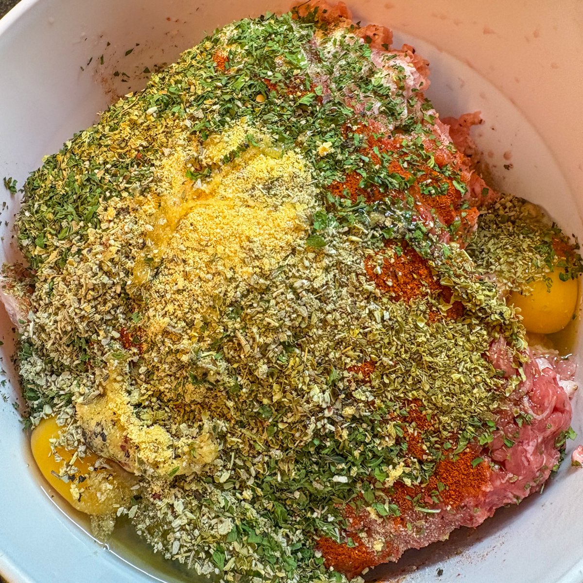Meatball mixture for baked Mediterranean meatballs in a white bowl, with pork and beef mince, eggs, paprika, garlic powder, onion powder and dried oregano, thyme, sage and parsley before mixing.