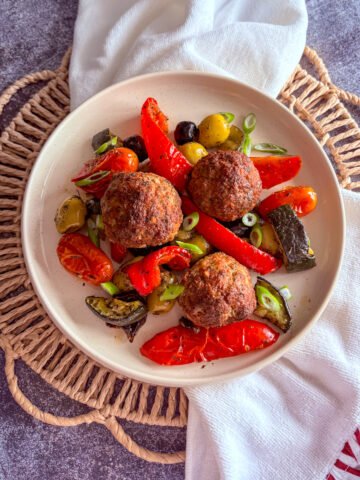 One tray meatballs and vegetables in a speckled cream bowl, top-down view of three browned meatballs with roasted peppers, olives, tomatoes and courgette on a rattan placemat with a white and red tea towel.