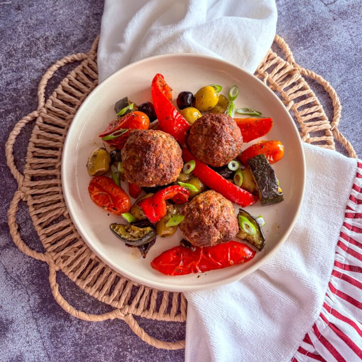 One tray meatballs and vegetables in a speckled cream bowl, top-down view of three browned meatballs with roasted peppers, olives, tomatoes and courgette on a rattan placemat with a white and red tea towel.