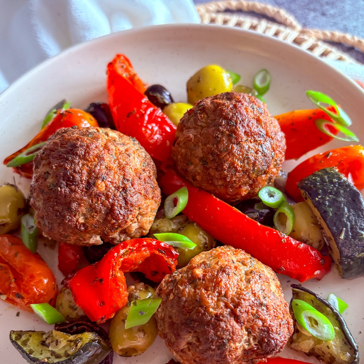 Baked Mediterranean meatballs close-up on a white plate, showing the browned, herb-speckled crust of three meatballs with roasted red pepper strips, a soft green olive, a courgette slice and fresh spring onion rings.